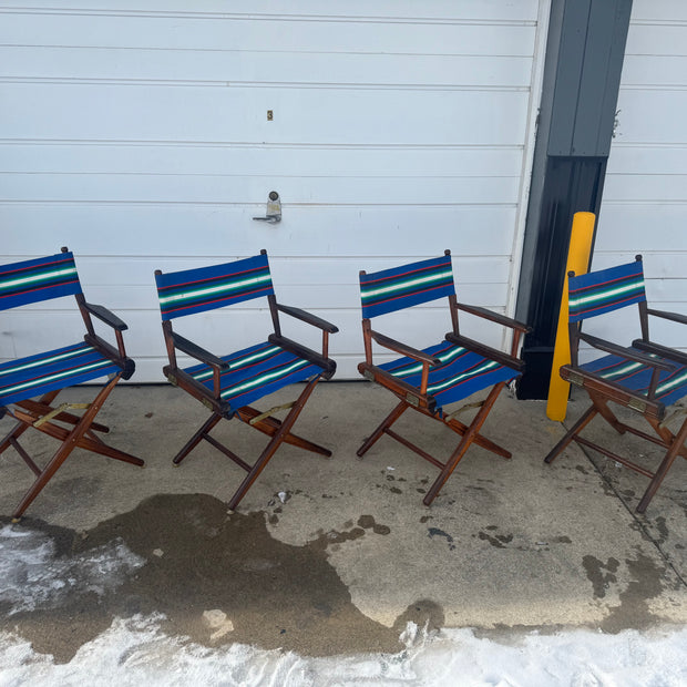 Four vintage canvas and wood folding captain's chairs placed outdoors, featuring a blue and white striped seat and backrest with a brown wood frame.