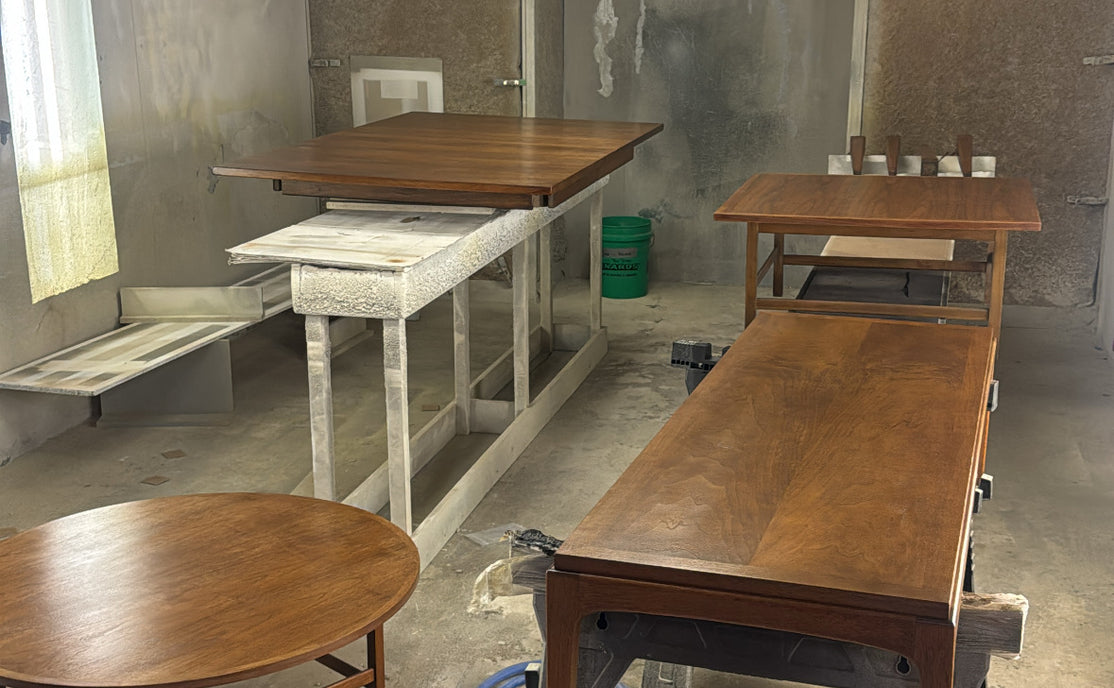 Several wooden tables and workbenches in a dusty workshop or furniture finishing space, with concrete floors, fluorescent ceiling lights, and a blue air hose in the foreground.