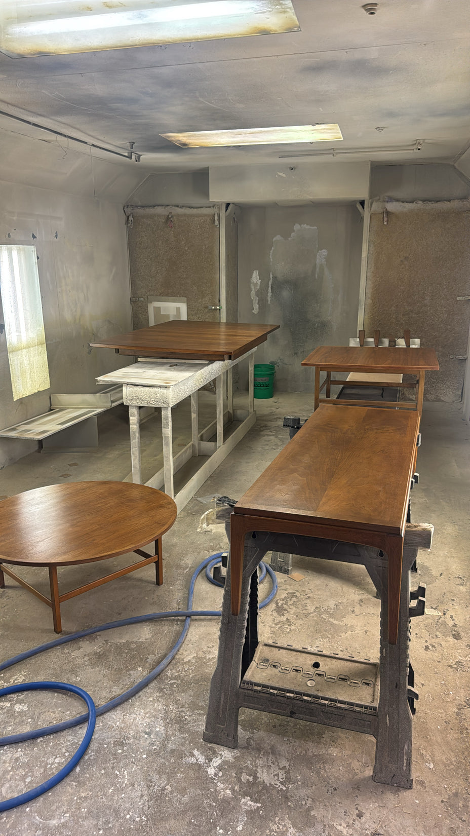 Several wooden tables and workbenches in a dusty workshop or furniture finishing space, with concrete floors, fluorescent ceiling lights, and a blue air hose in the foreground.