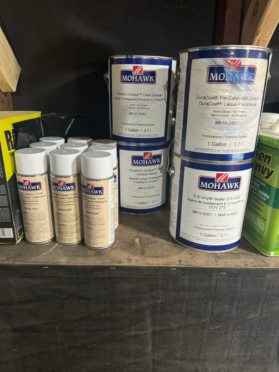 Several cans of Mohawk wood finish and stain products arranged on a wooden shelf, including stacked blue-and-white paint cans and smaller beige spray cans, with a dark wall behind them.