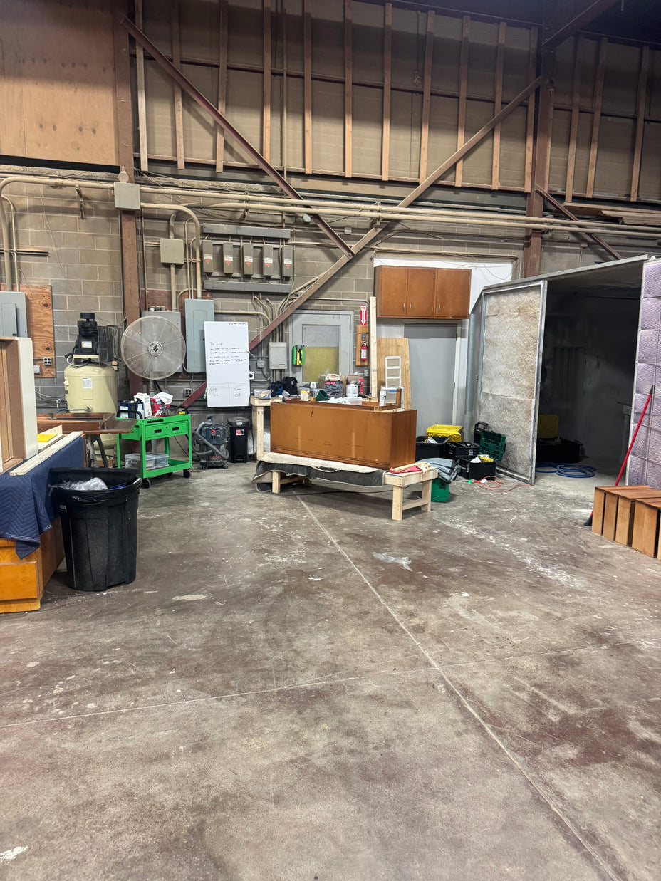 A large, cluttered warehouse or workshop space with a concrete floor, exposed wooden ceiling beams, and various pieces of equipment, furniture, and materials scattered throughout the room.