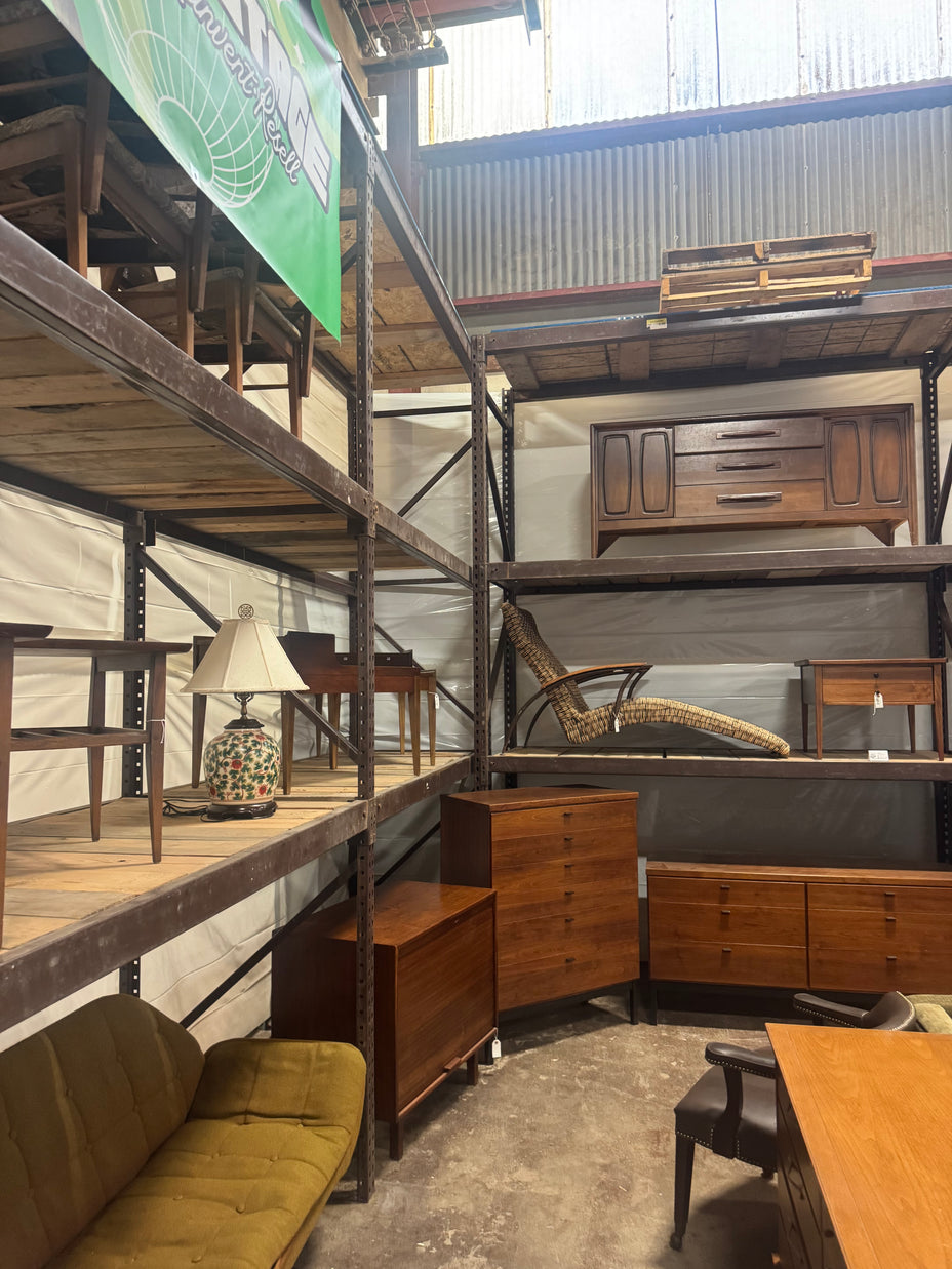 A warehouse-style showroom filled with vintage wooden furniture, including dressers, a desk, shelving, a chair, and a green upholstered bench, with metal support racks throughout.