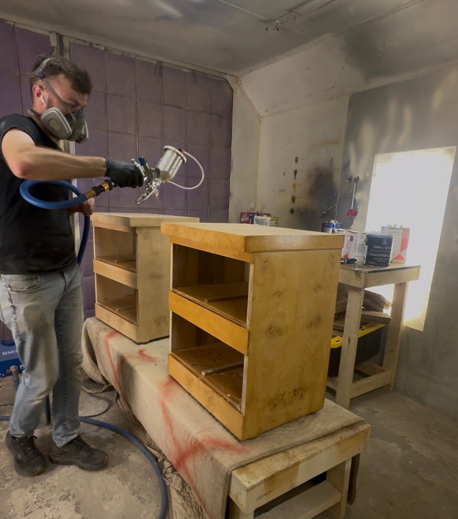 A man wearing a respirator mask and gloves uses a spray gun to apply finish to a light wood cabinet in a workshop setting.